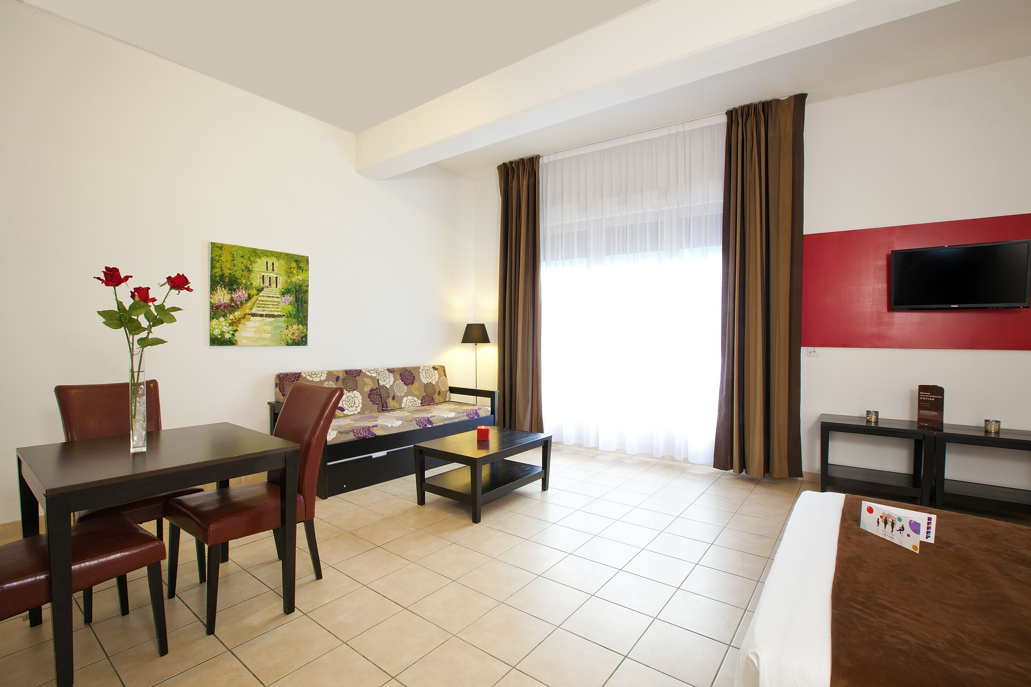 Una habitación de hotel con cama, televisor, sofá, mesa de comedor con sillas, un jarrón con flores y un cuadro en la pared.