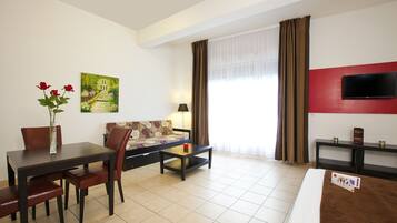 Una habitación de hotel con cama, televisor, sofá, mesa de comedor con sillas, un jarrón con flores y un cuadro en la pared.