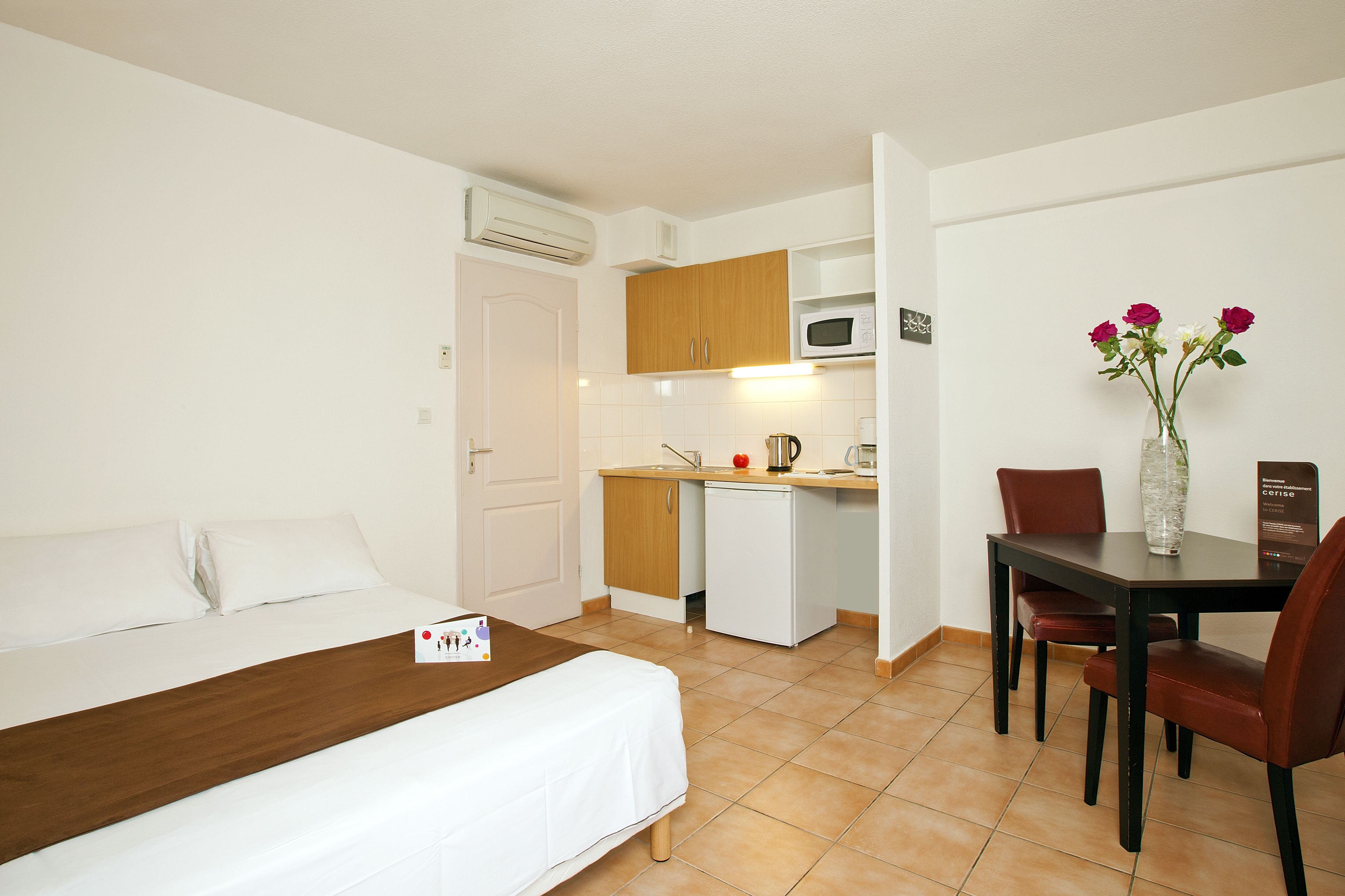 Une chambre d’hôtel comprenant un lit, une petite kitchenette, une table à manger avec des chaises et un vase avec des fleurs.