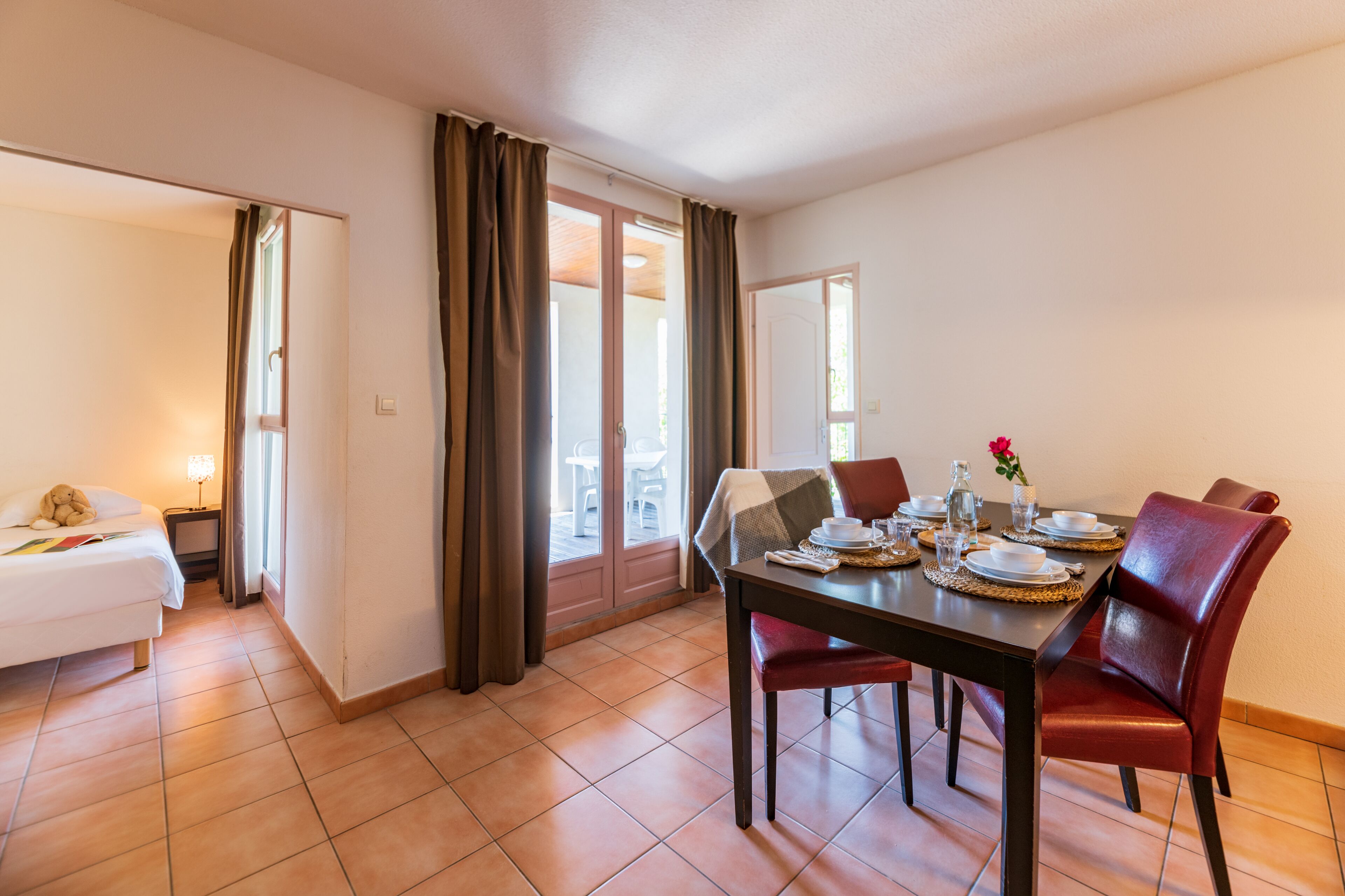 A hotel room with a dining area featuring a table set for four, a bed, and a view through a sliding door.