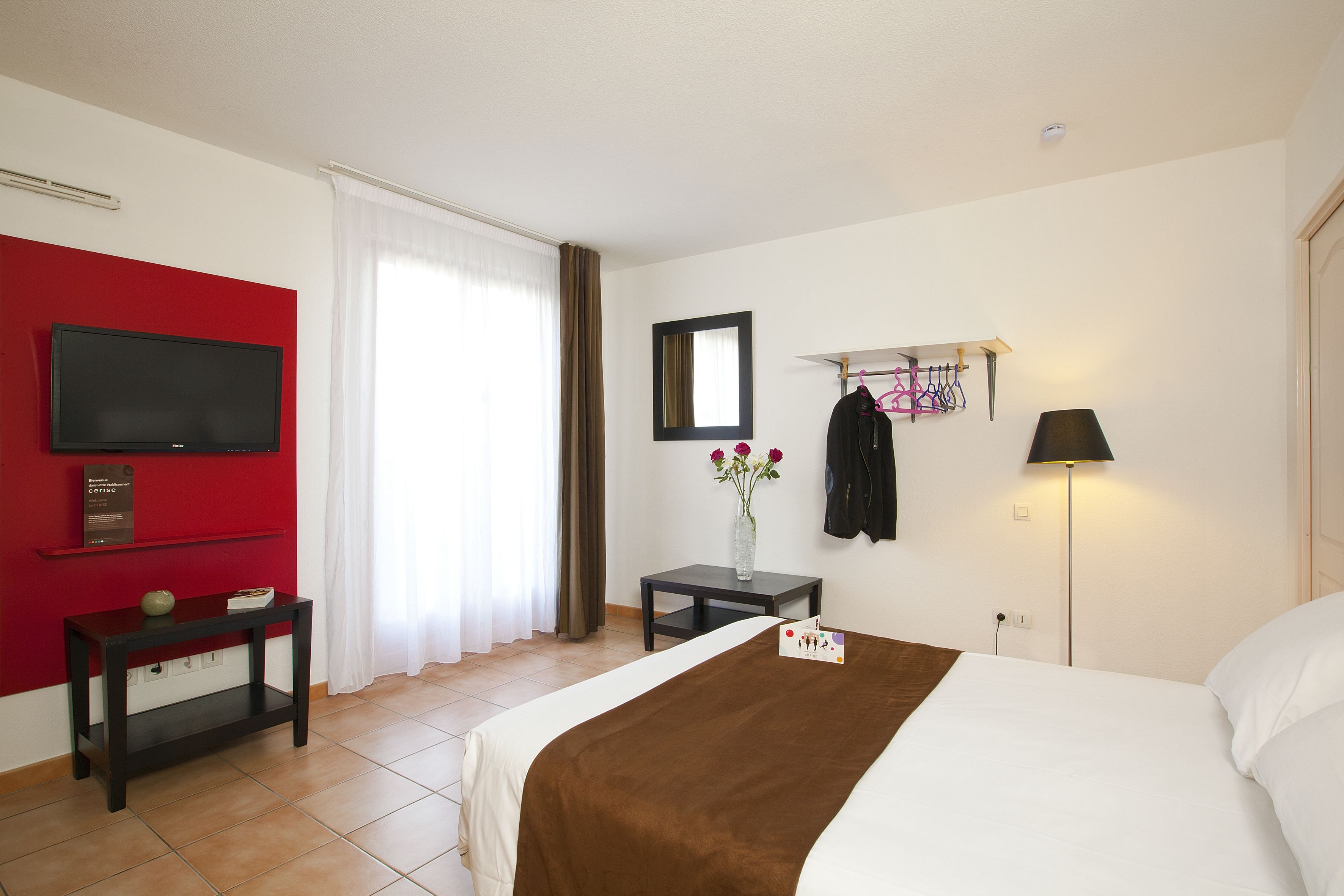 A hotel room with a bed, a TV mounted on the wall, a small table with a vase of flowers, and a coat rack with hangers.