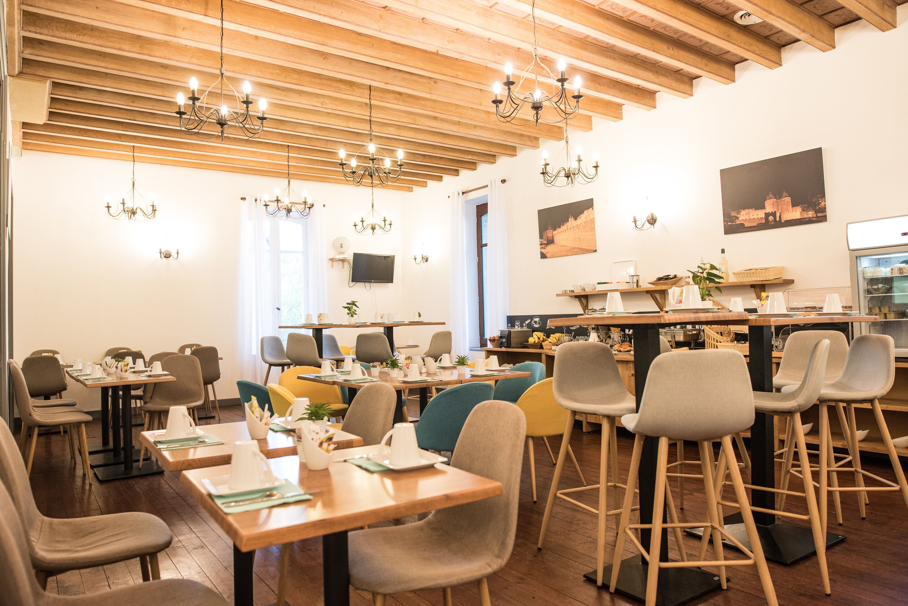 A restaurant with wooden ceiling beams, multiple tables and chairs, and hanging chandeliers.