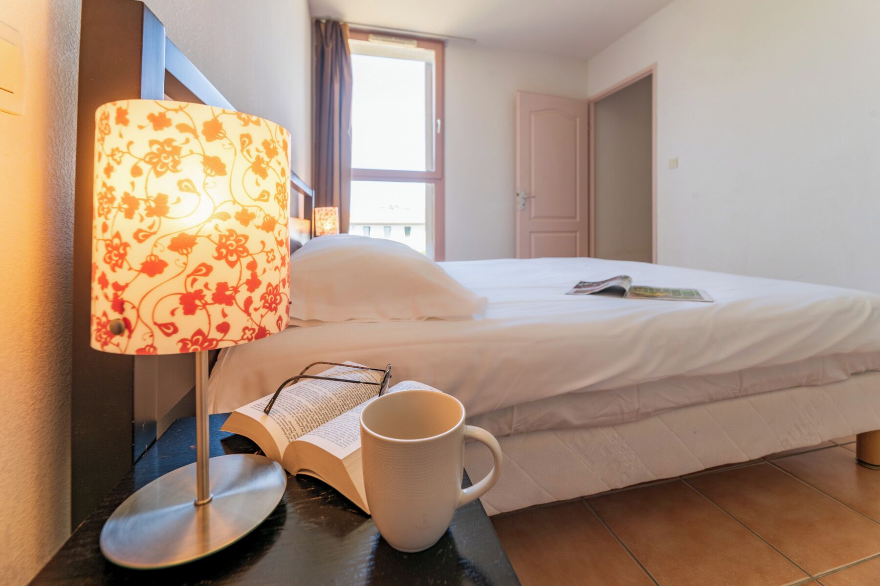 Une chambre d’hôtel avec un lit, une table de chevet avec une lampe, un livre et une tasse.