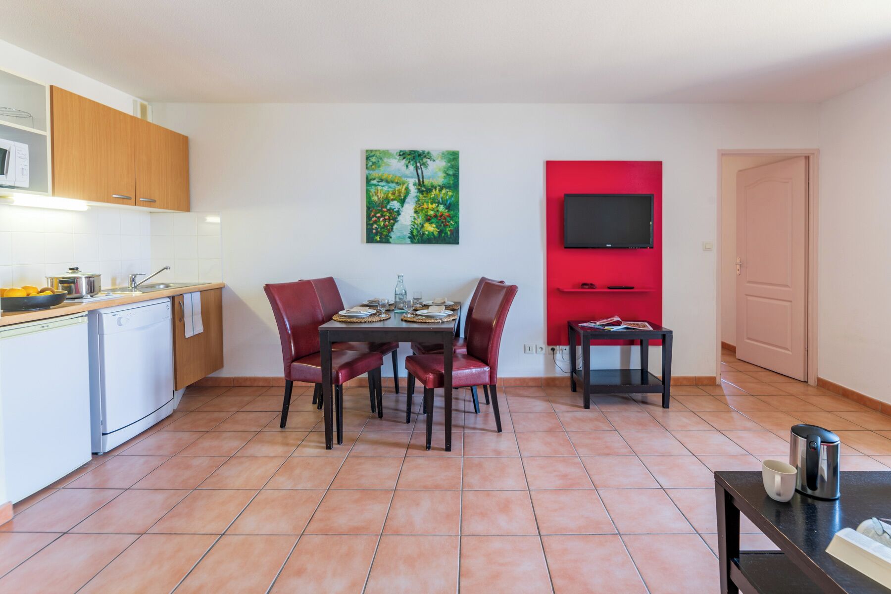 A compact kitchen with a dining area, a red accent wall, and a flat-screen TV mounted on it.