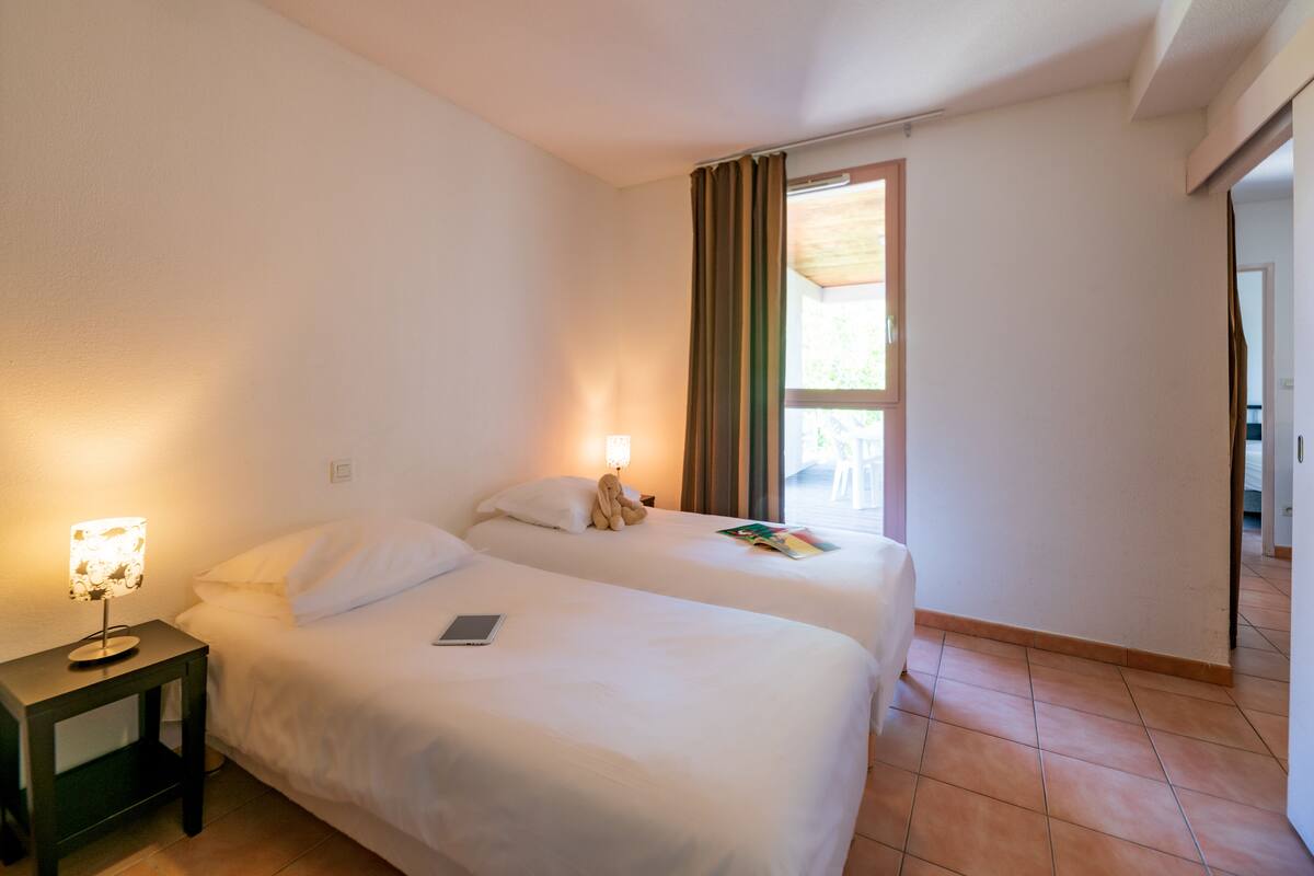 Ein Hotelzimmer mit zwei Betten, einem Nachttisch mit Lampe, einem Fenster mit Vorhängen und Blick ins Freie.