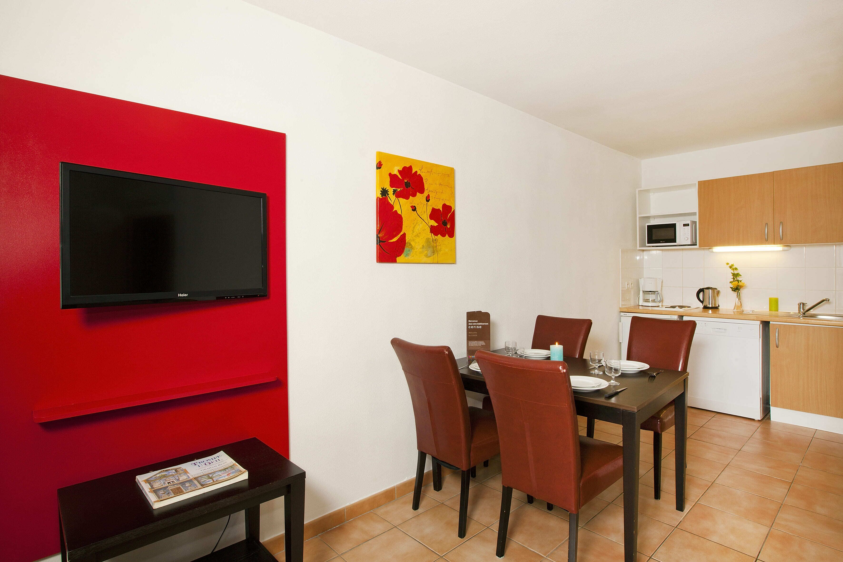 A dining area with a TV mounted on a red wall, a table set with chairs, and a kitchen area with a microwave and cabinets.