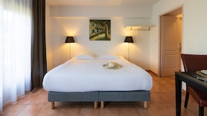 Una cama bien hecha con edredón blanco y almohadas, dos lámparas de noche, un cuadro en la pared y una puerta a la derecha.