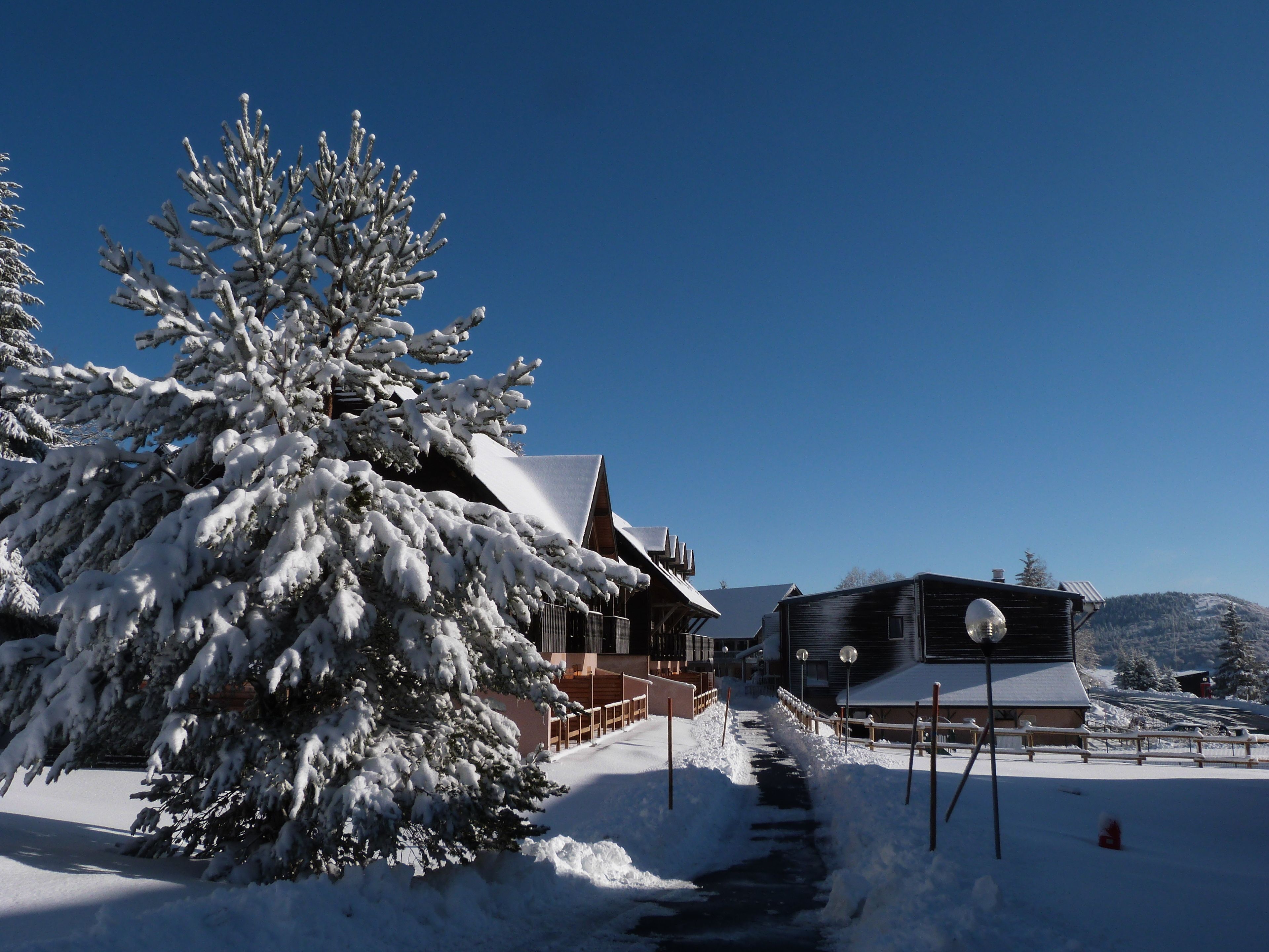 Un paisaje nevado con un edificio que tiene una terraza de madera y un tejado cubierto de nieve.