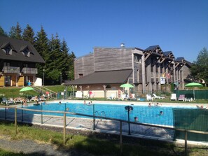 A swimming pool area with lounge chairs and umbrellas, surrounded by wooden buildings and trees. - Résidence Le Bois de la Reine (Besse-et-Saint-Anastaise)