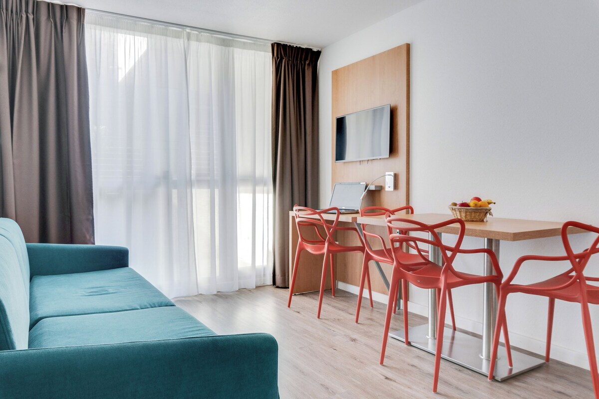 A modern living room with a teal sofa, a dining area with red chairs, and a wooden table with a basket of fruit.