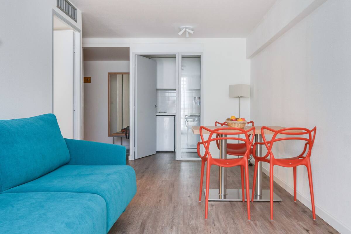 Una sala de estar moderna con un sofá color turquesa, una mesa de comedor con cuatro sillas naranjas, y una cocina visible a través de una puerta abierta.