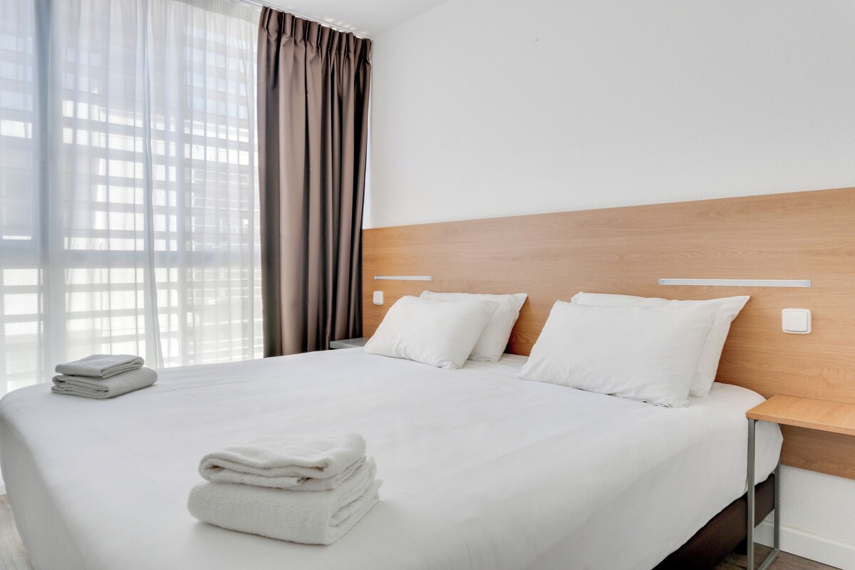 Une chambre d’hôtel avec un grand lit, du linge de lit blanc, une tête de lit en bois et une vue à travers des rideaux transparents.