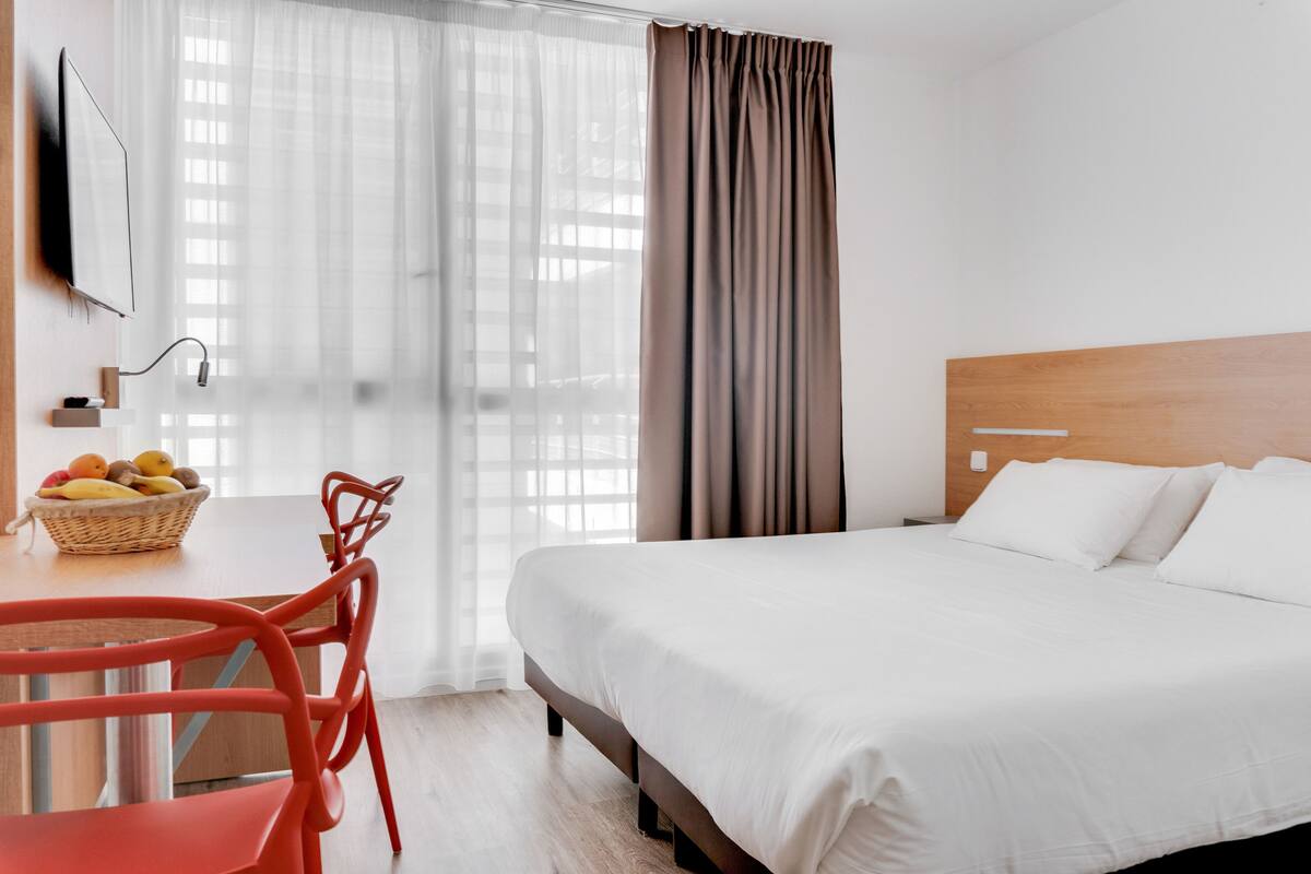 Habitación de hotel con una cama, un escritorio con una cesta de frutas, una silla y un televisor montado en la pared.