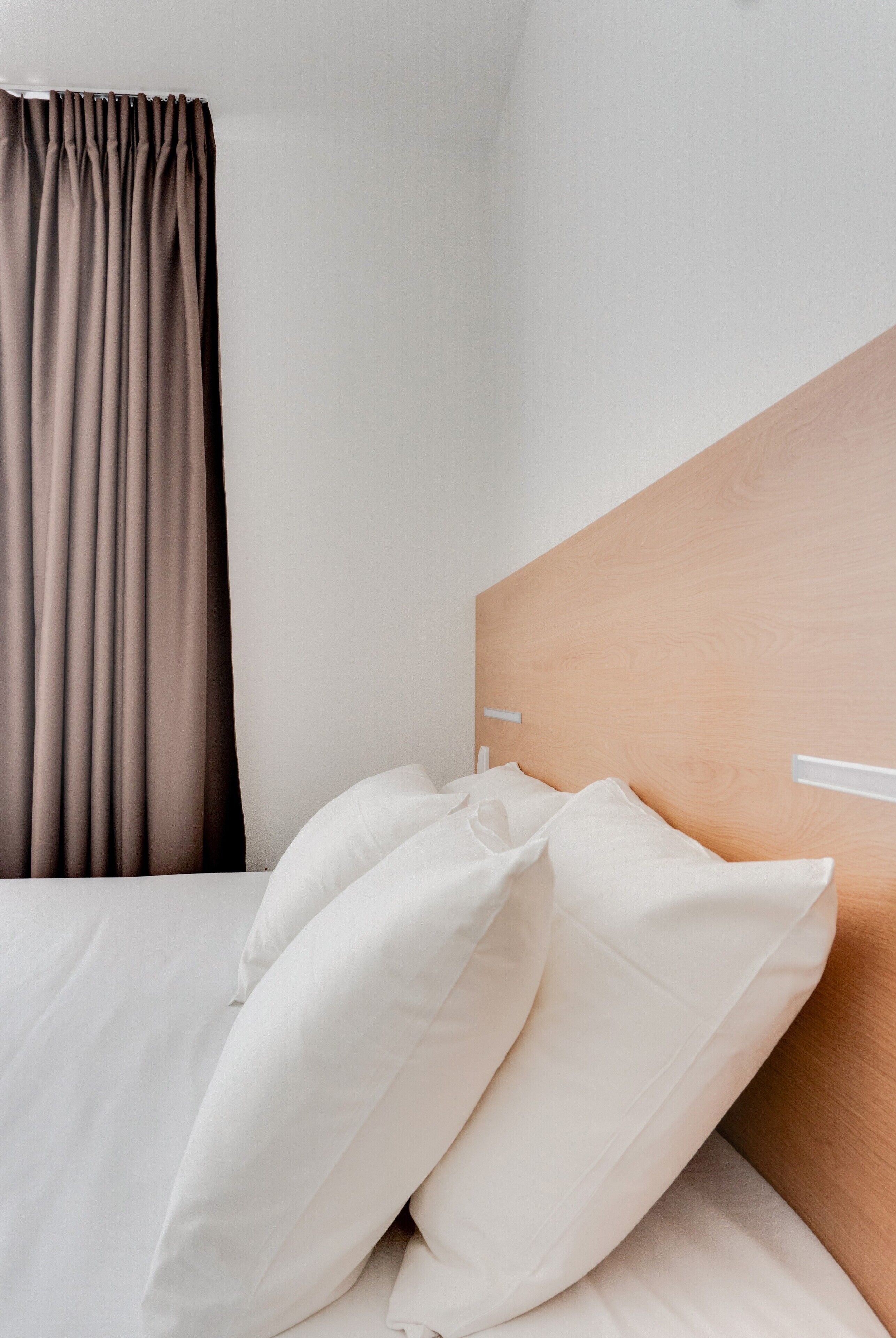 A hotel room with a wooden headboard, white bedding, and a brown curtain.