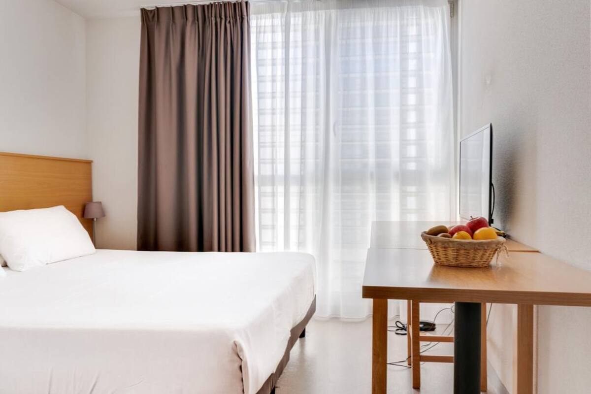 Habitación de hotel con cama, una mesa con frutero, televisor y cortinas.