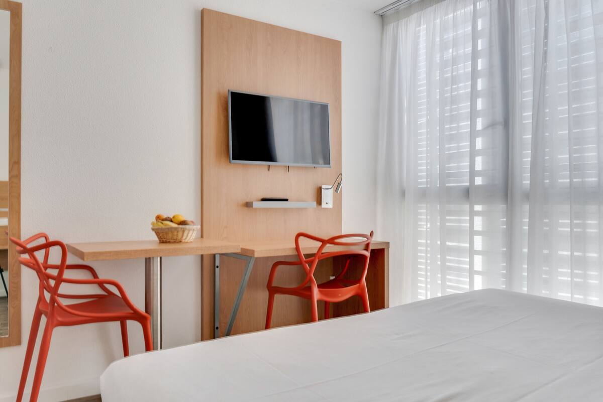 Habitación de hotel con cama, escritorio y dos sillas rojas, televisor montado en la pared y ventana con cortinas translúcidas.