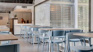 Un espace de restauration moderne avec des chaises bleu clair et des tables en bois, de grandes fenêtres équipées de stores et un design épuré et minimaliste.