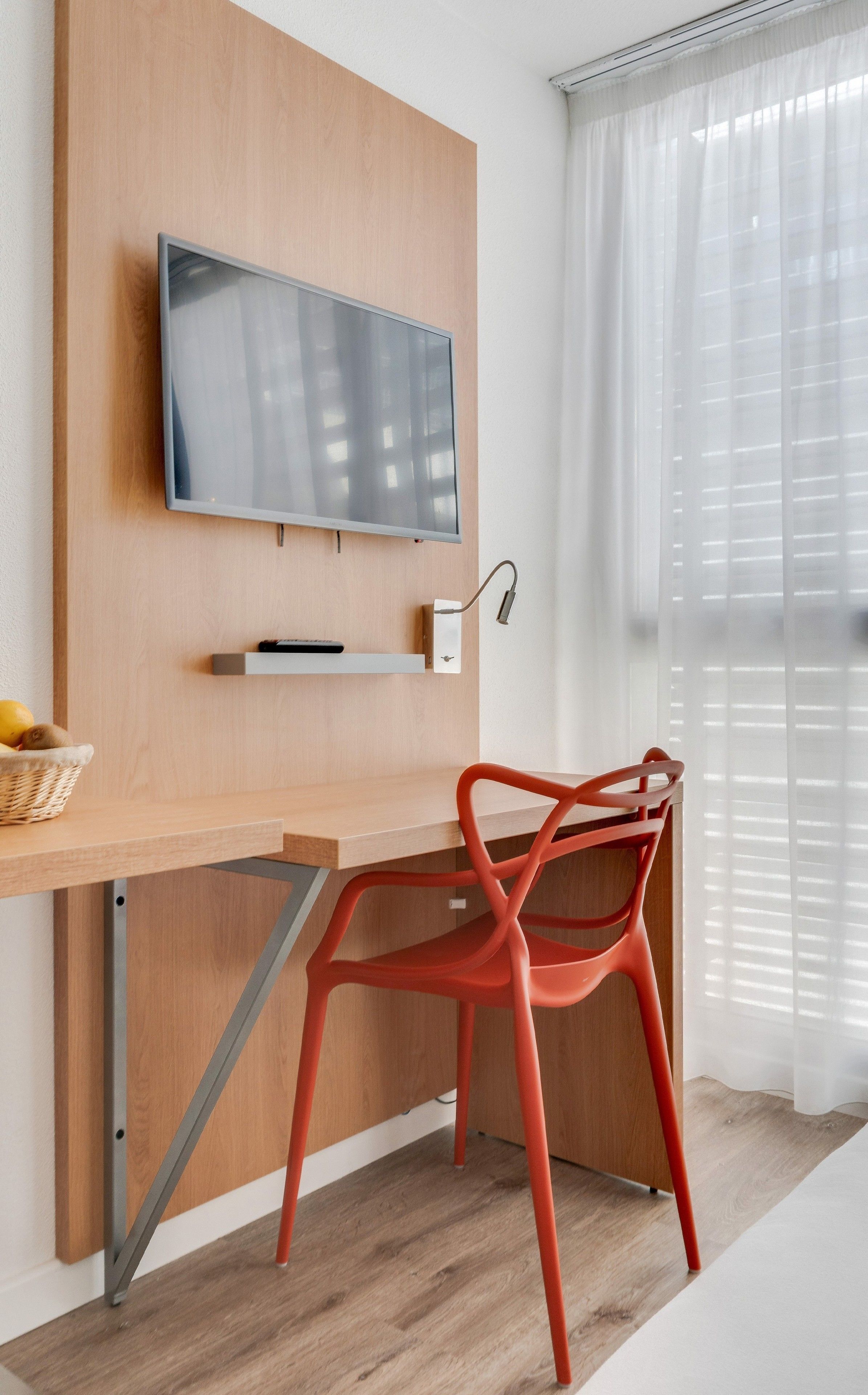 A modern room with a wooden desk, a red chair, a mounted TV, and a window with sheer curtains.