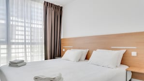 Une chambre d’hôtel avec un grand lit, du linge de lit blanc, une tête de lit en bois et une vue à travers des rideaux transparents.
