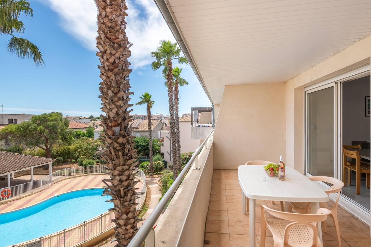 Un balcon donnant sur une piscine, des palmiers et une table avec des chaises.