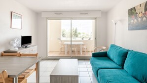 A modern living room with a teal sofa, a dining area with a wooden table and chairs, a TV on a cabinet, and a large sliding glass door leading to an outdoor patio. - Résidence Residhotel Le Mayflower (Le Grau-Du-Roi)