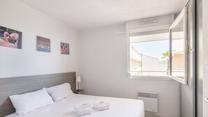 A hotel room with a bed, white linens, a nightstand with a lamp, and wall art. - Résidence Residhotel Le Mayflower (Le Grau-Du-Roi)