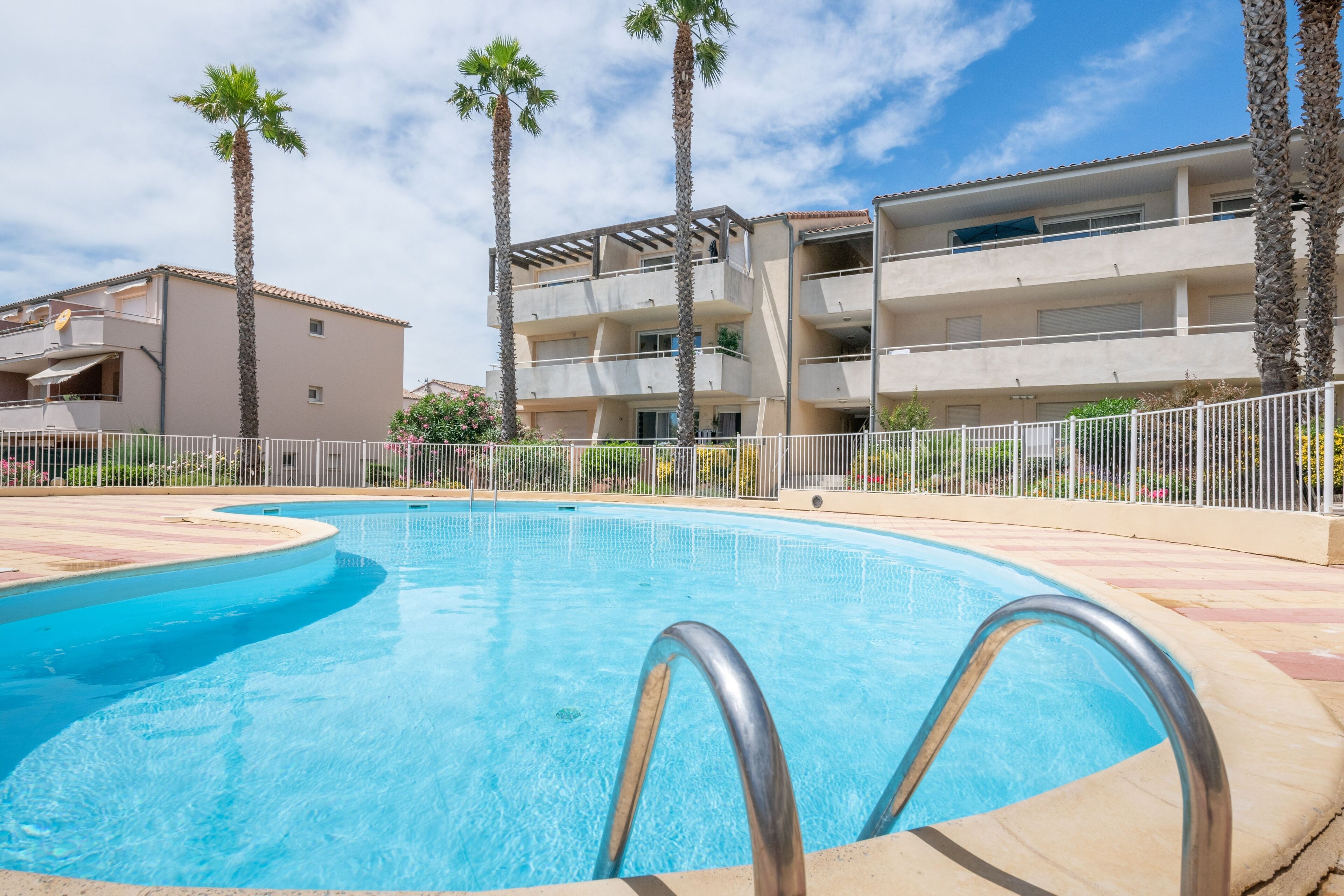 A swimming pool with a metal ladder, surrounded by a white fence, palm trees, and a multi-story building with balconies.