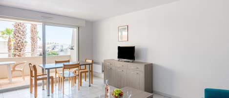 Una sala de estar moderna con comedor, un televisor de pantalla plana montado en la pared y un balcón con vistas a palmeras y edificios.