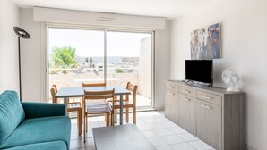 A modern living room with a sofa, a dining table with chairs, a TV on a cabinet, and a balcony with a city view. - Résidence Residhotel Le Mayflower (Le Grau-Du-Roi)