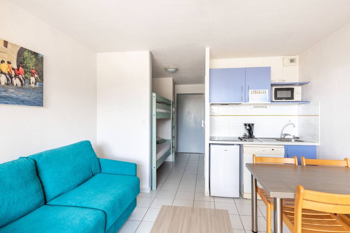 Une pièce de taille modeste, avec un canapé de couleur turquoise, une table à manger en bois et un coin cuisine équipé d’un four à micro-ondes et d’un évier.