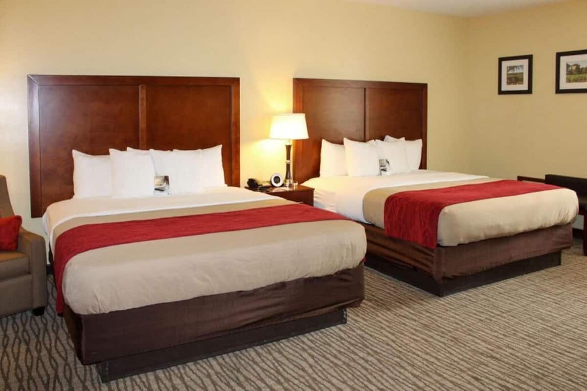 A hotel room with two beds, a nightstand with a lamp, and a chair.