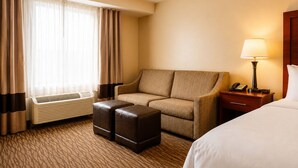 A hotel room with a sofa, an ottoman, a bed, a nightstand with a lamp, a telephone, and a window with curtains.