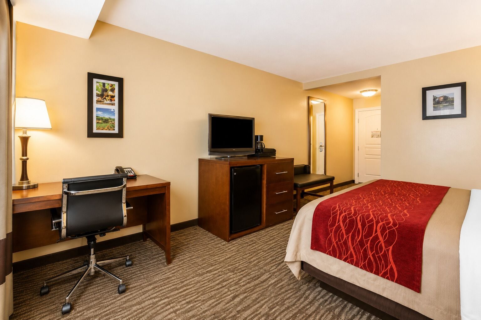 A hotel room with a desk, chair, television, bed, and a door leading to another room.