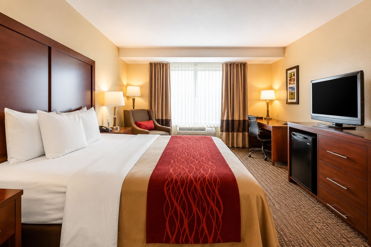 A hotel room with a large bed, a desk with a television, a chair, and a desk lamp.