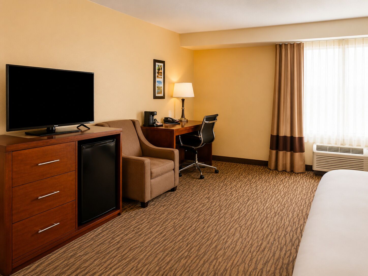 Habitación de hotel con televisión de pantalla plana, escritorio con lámpara, una silla, una cama y aire acondicionado.