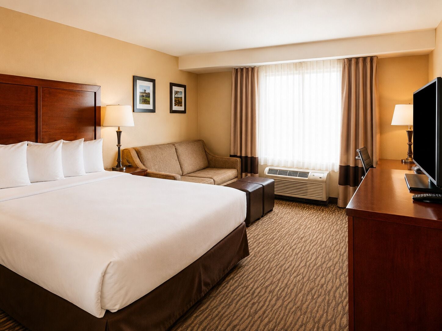 A hotel room with a large bed, a sofa, a desk with a television, and an air conditioning unit.