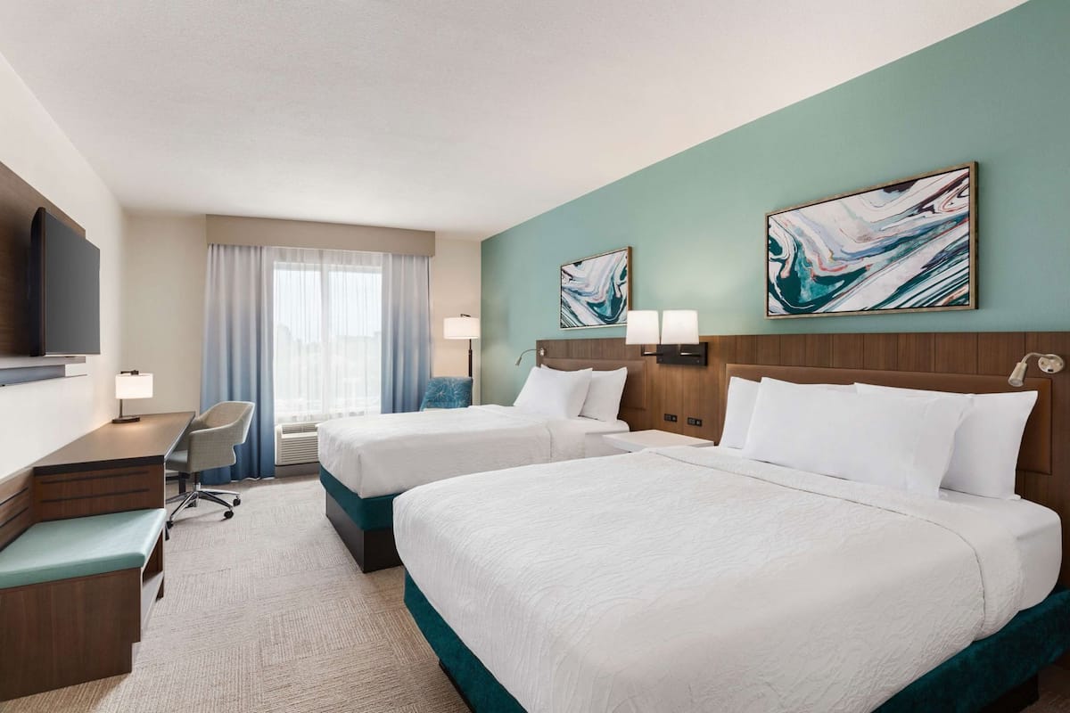 A hotel room with two beds, a desk, a chair, a TV, and artwork on the walls.
