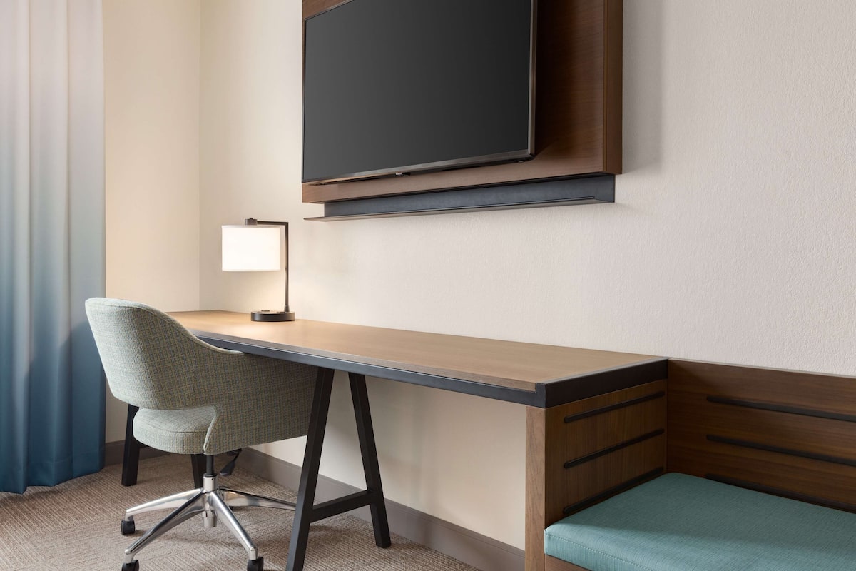 A hotel room with a desk, chair, TV, and a small bed.