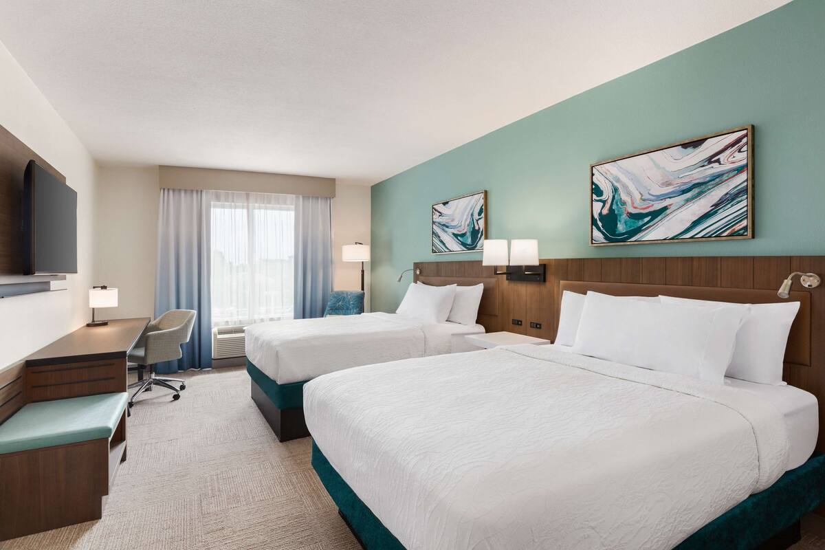 A hotel room with two beds, a desk, a chair, and a TV.