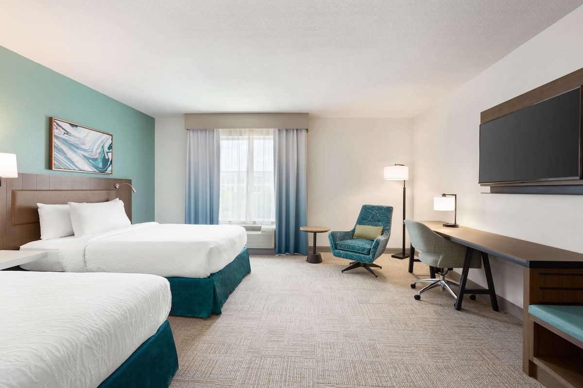 A hotel room with two beds, a desk, a chair, a TV, and a window with curtains.