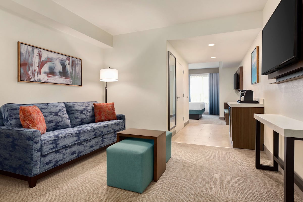 A modern hotel room with a blue sofa, teal ottoman, a desk, and a flat-screen TV mounted on the wall.