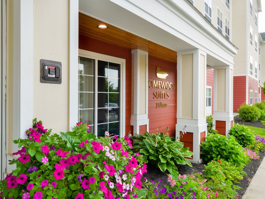 A hotel entrance with a sign reading "OMEWOOD SUITES" and a well-maintained garden with various flowers.