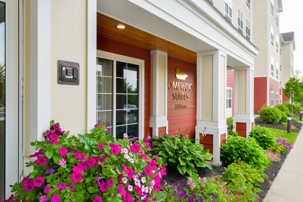 A hotel entrance with a sign reading "OMEWOOD SUITES" and a well-maintained garden with various flowers.
