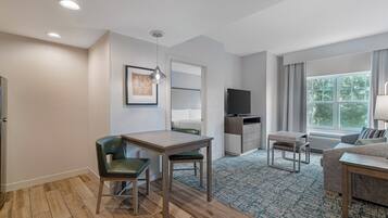 A modern hotel room with a wooden floor, a desk with a chair, a sofa, a television, and a window with curtains.