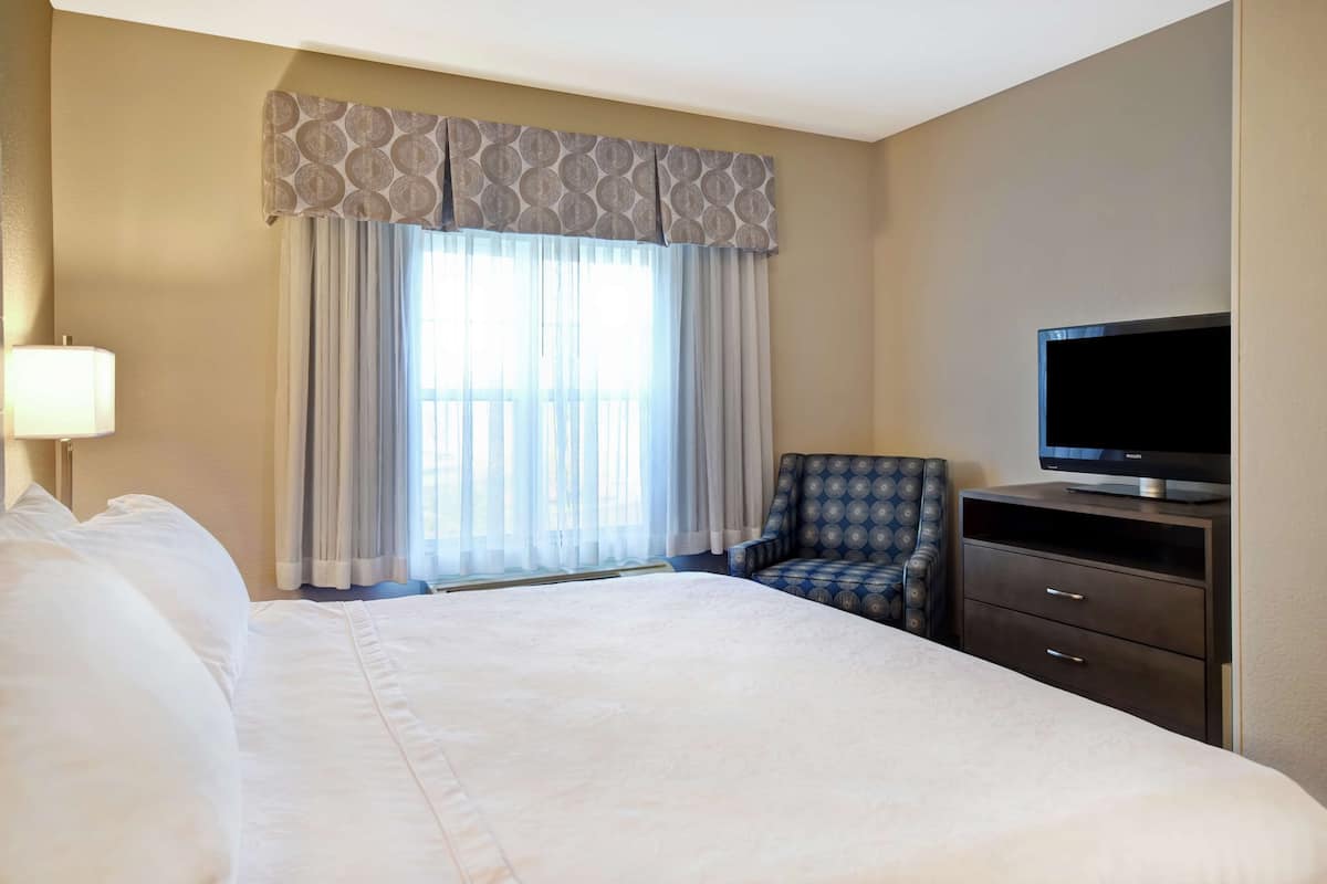 Een hotelkamer met een groot bed, een televisie op een dressoir, een stoel en een raam met gordijnen.