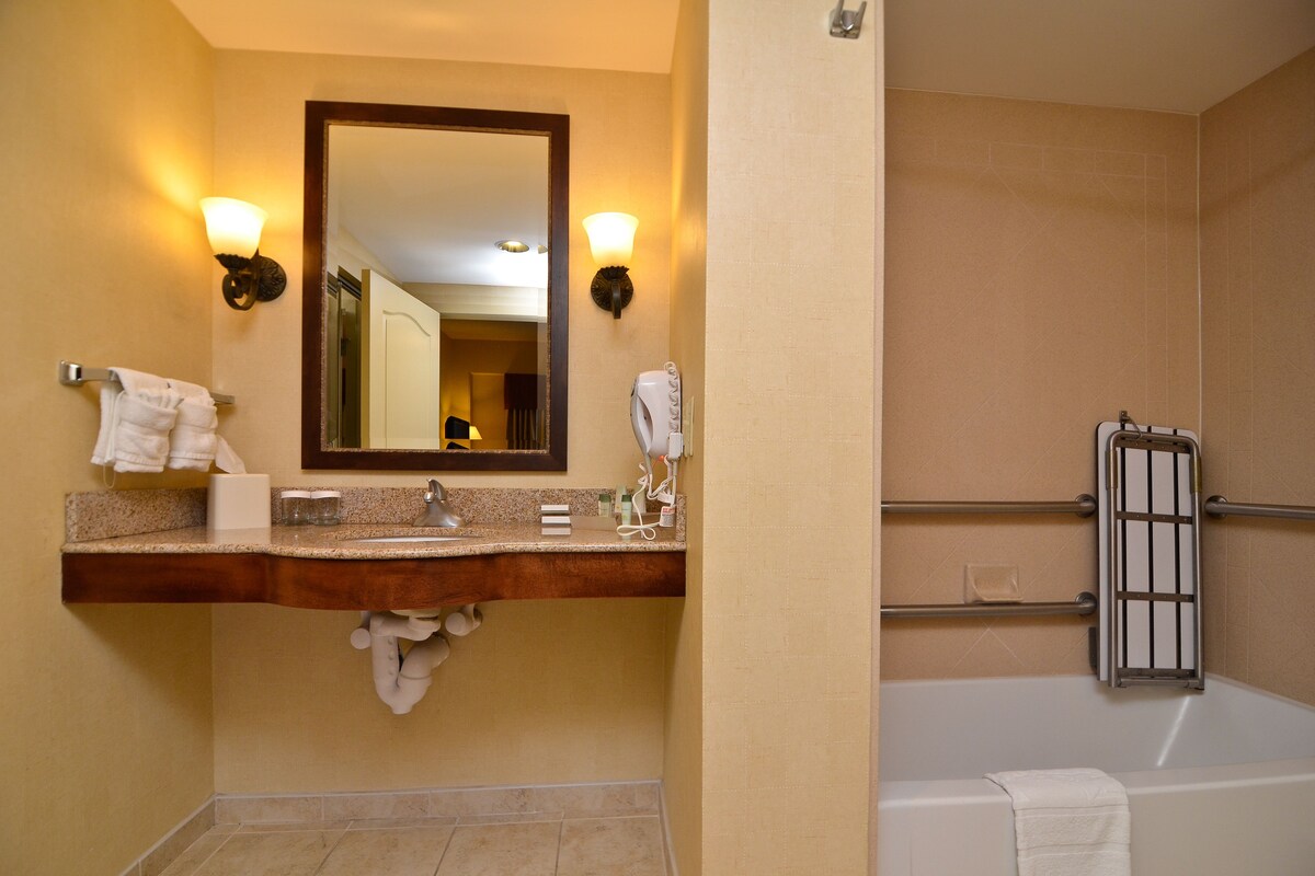 A bathroom with a wooden vanity, a large mirror, two wall-mounted lights, a towel rack, a hairdryer, and a bathtub with a safety bar.