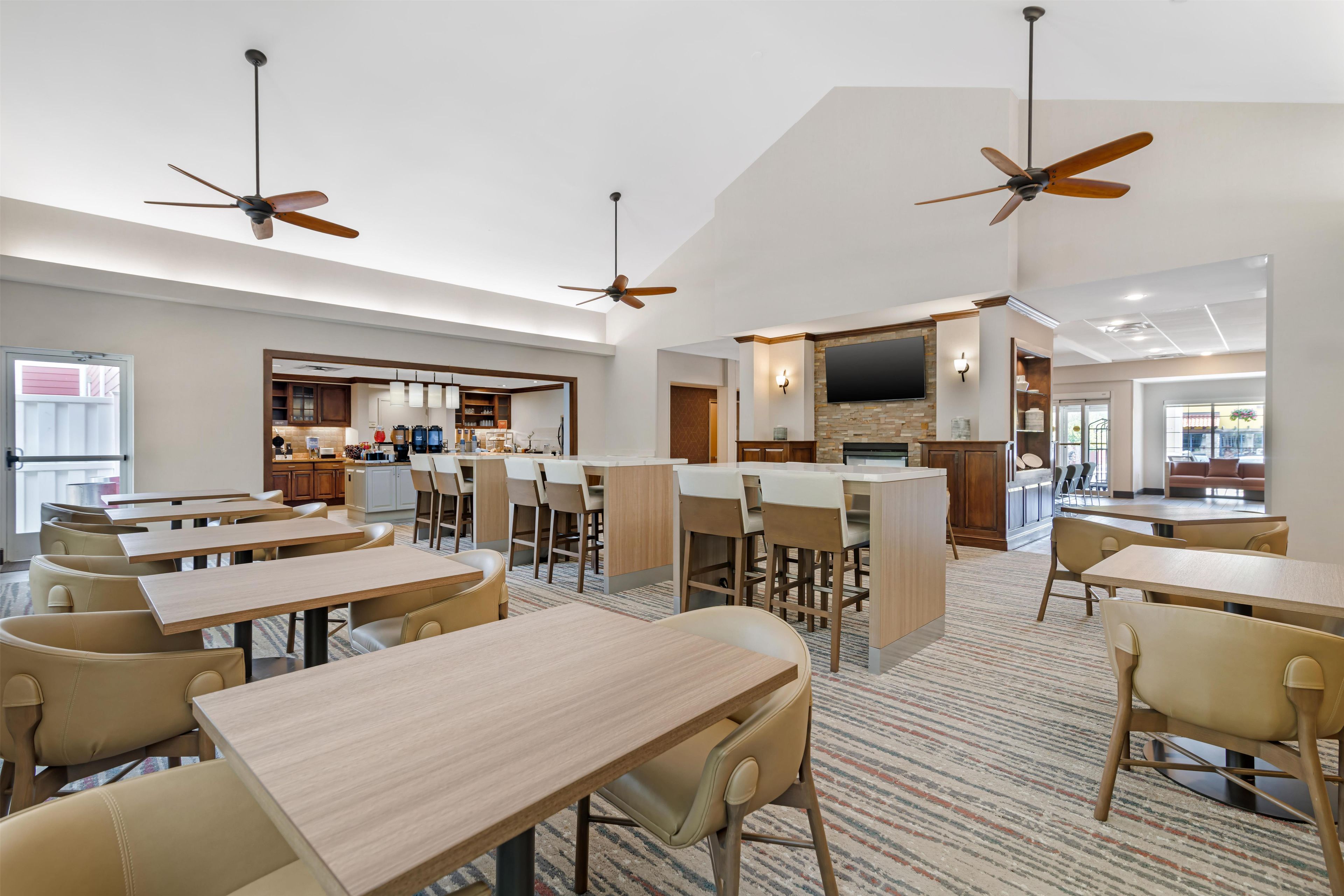 A hotel dining area with multiple tables and chairs, ceiling fans, a kitchen area, and a television mounted on the wall.