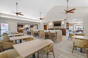 Een restaurantruimte in een hotel met meerdere tafels en stoelen, plafondventilatoren, een keukenhoek en een televisie aan de muur.