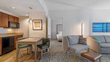 A modern hotel room with a kitchenette, a dining area, a living room with a sofa and a painting, and a bedroom with a bed and a nightstand.