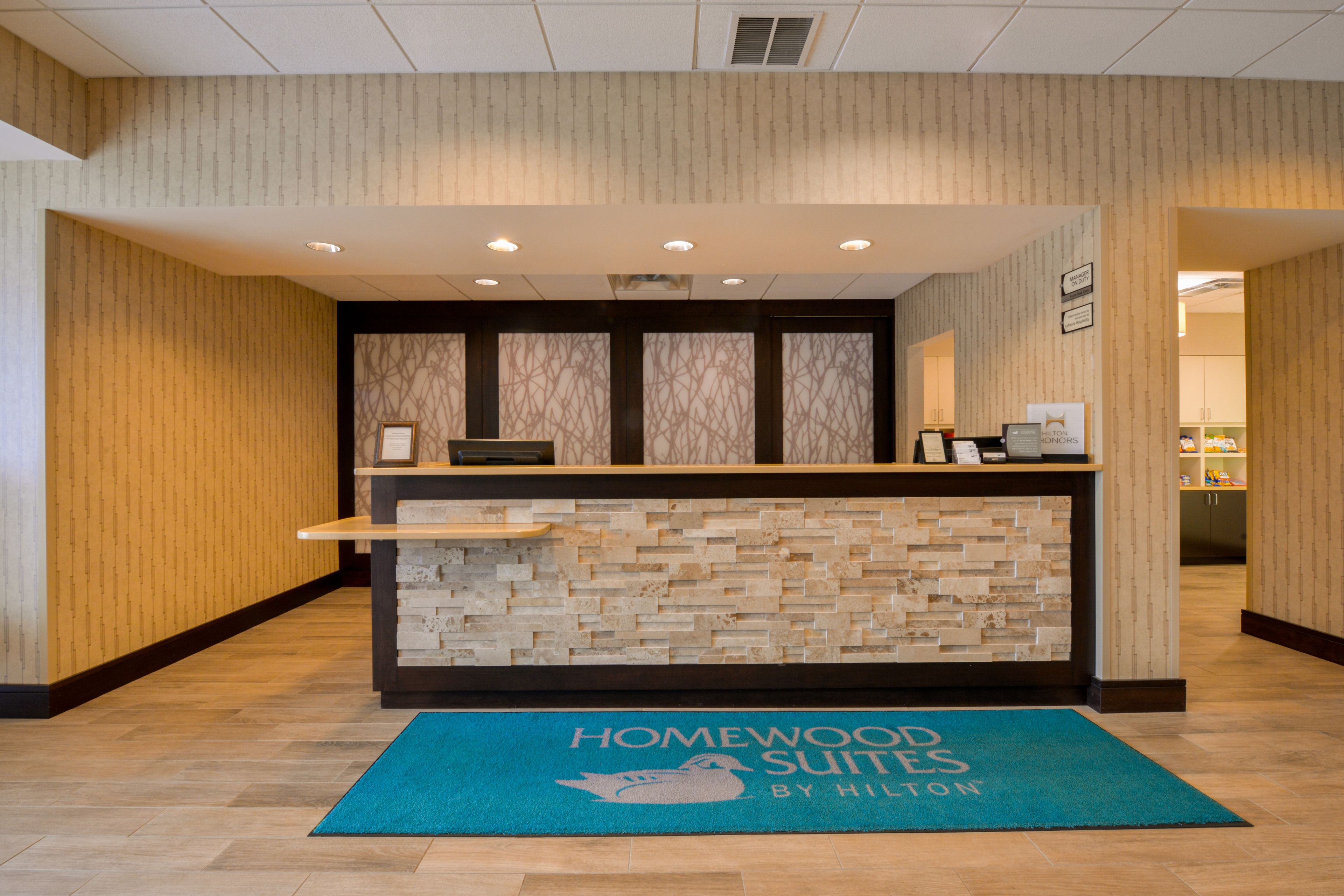 A reception desk with a wooden and brick design, a blue mat with "Homewood Suites by Hilton" text, and a reception area with a patterned wall and a framed picture.